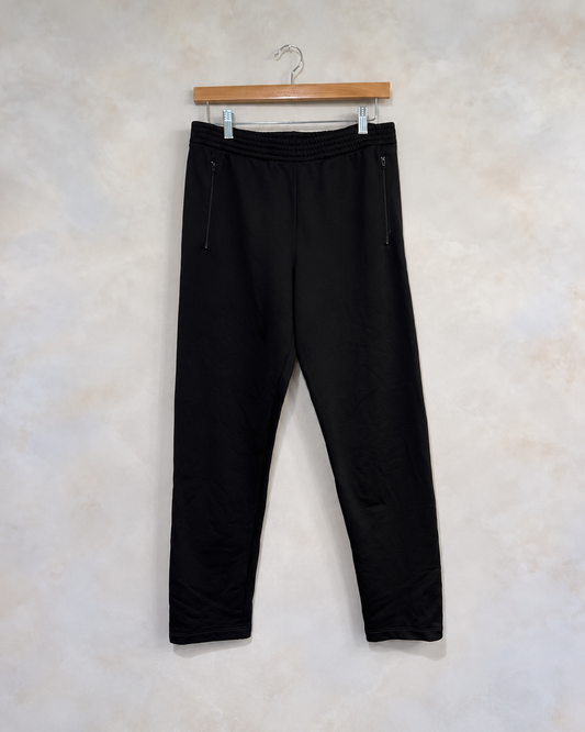 Balenciaga black track joggers with zip pockets on wooden clip hanger against light gray beige wall
