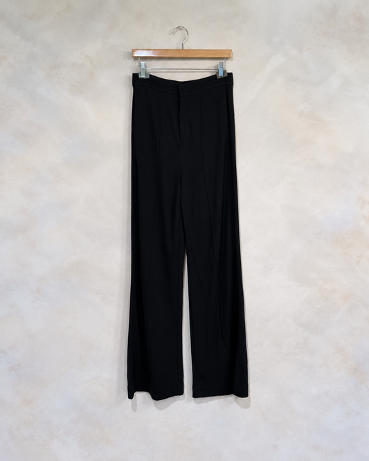 Rag & Bone black wide leg jersey trousers on wooden clip hanger against light gray beige wall